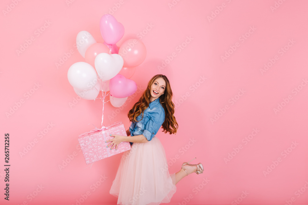 Excited long-haired girl in lush midi skirt standing on one leg, holding cute birthday present. Charming curly young woman in stylish outfit going to friend's party with gift and colorful balloons - obrazy, fototapety, plakaty 