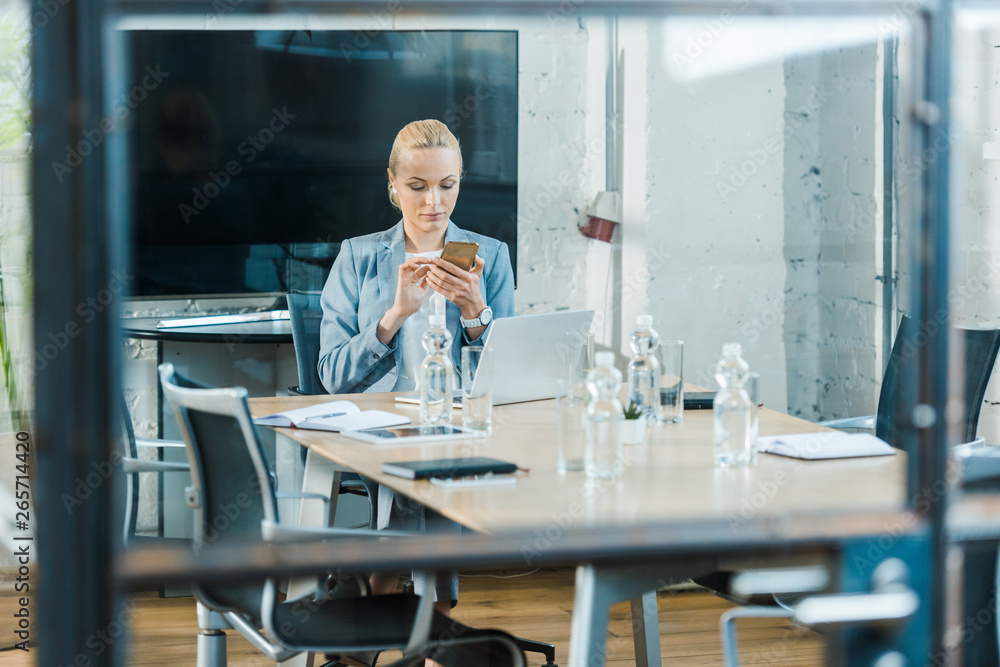 selective focus of attractive blonde businesswoman sitting in conference room and using smartphone