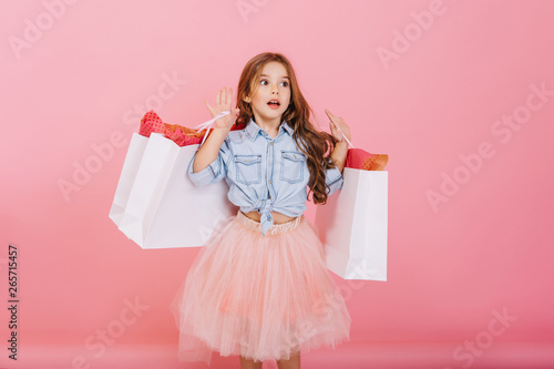 Amazing little astonished  girl in tulle skirt expressing to side with white packages in hands isolated on pink background. Young shopper with presents, shopping in childhood