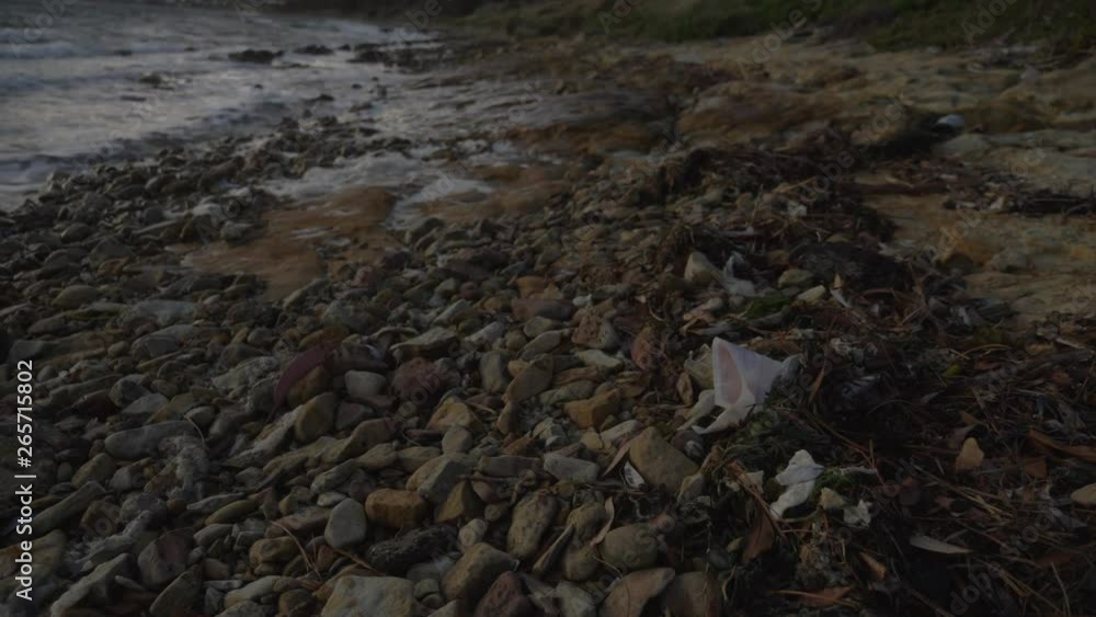 Piece of white plastic rubbish sits among rocks on coastal foreshore on windy day while waves roll in. Piece of white plastic rubbish sits among rocks on coastal foreshore on windy day while waves roll in.