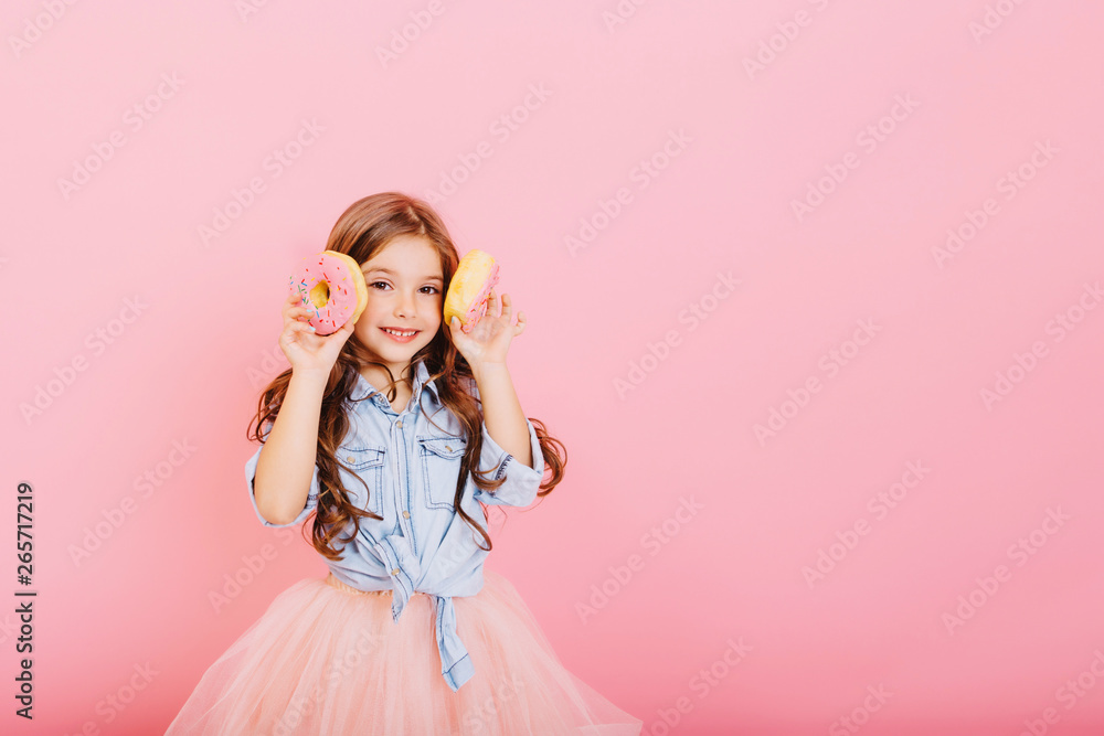 Cute little girl with long brunette hair having fun to camera with donuts isolated on pink background. Charming fashionable princess in tulle skirt expressing positivity. Place for text