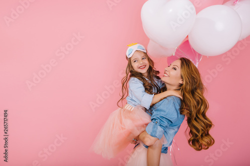 Adorable laughing birthday girl with colorful balloons embracing her young smiling mom after funny event. Attractive mother posing with pretty daughter in mask at party isolated on pink background