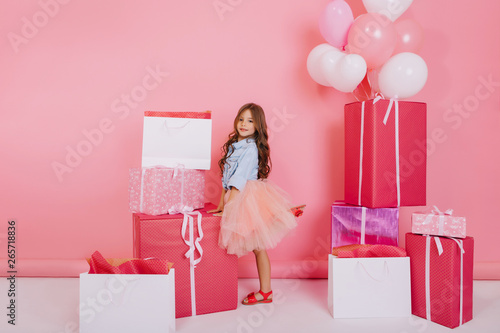 Friendly little girl with long curly hair in pink tulle skirt smiling to camera suround colorful big giftboxes, balloons on pink background. Happy childhood, birthday mood of cute princess photo