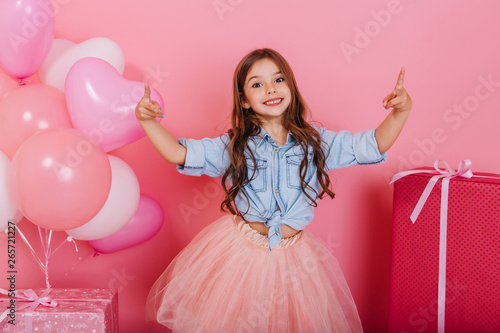 Expressing true positive emotions to camera of joyful happy kid suround balloons and presents isolated on pink background. Charming little princess in tulle skirt smiling, celebrating birthday photo