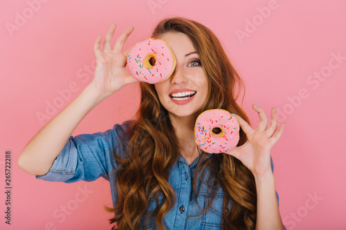 Fotografija Long-haired adorable girl in denim shirt plays with glazed donuts before tea-drinking with friends