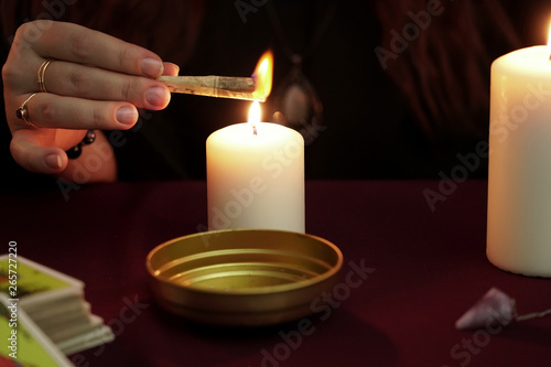 Witch is fortune teller in black mantle with rings conjures burning paper with a spell. Tarot cards, amethyst stones, white candles on dark background. Occult, esoteric, divination and wicca concept.