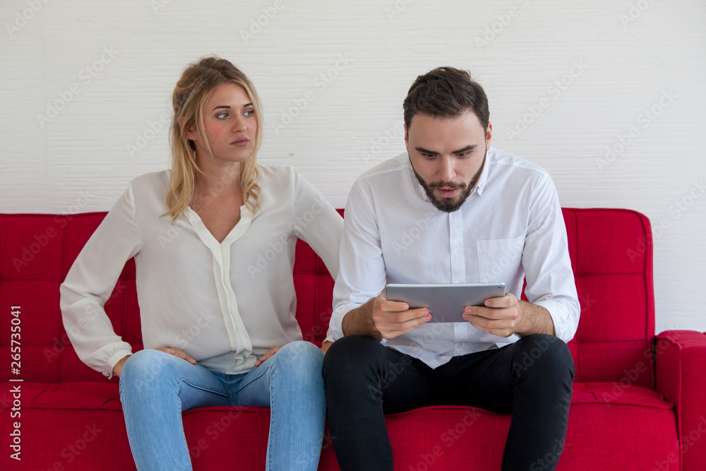 young man who is playing a game on a tablet. Young Couple Sitting On Sofa Using Digital Tablet. Women who are tired of men playing games young man who is playing a game on a tablet. Young Couple Sitting On Sofa Using Digital Tablet. Women who are tired of men playing games