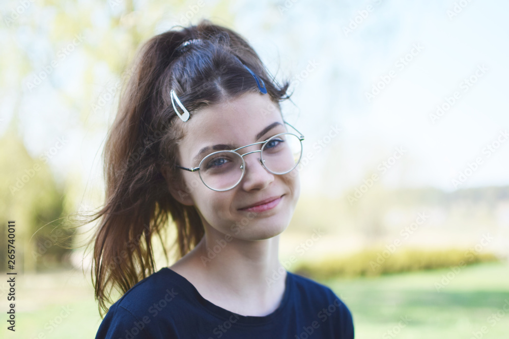 Portrait of pretty smiling teen girl wearing round glasses looking at  camera outdoors on blurred landscape background with copy space in spring  day. Stock Photo