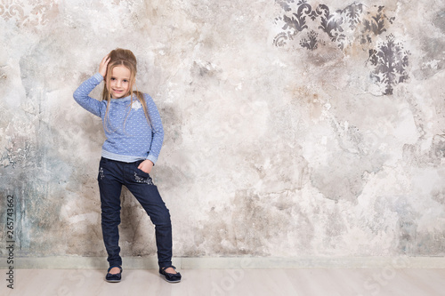 Portrait of a little attractive smiling girl in a blue sweater and pants with hair folded into her hair against a grunge wall background.