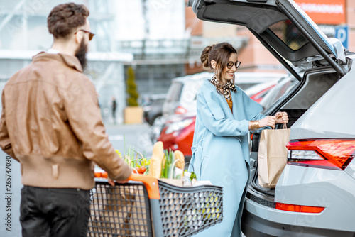 Young stylish couple with shopping cart full of fresh food, packing products into the car on the outdoor parking