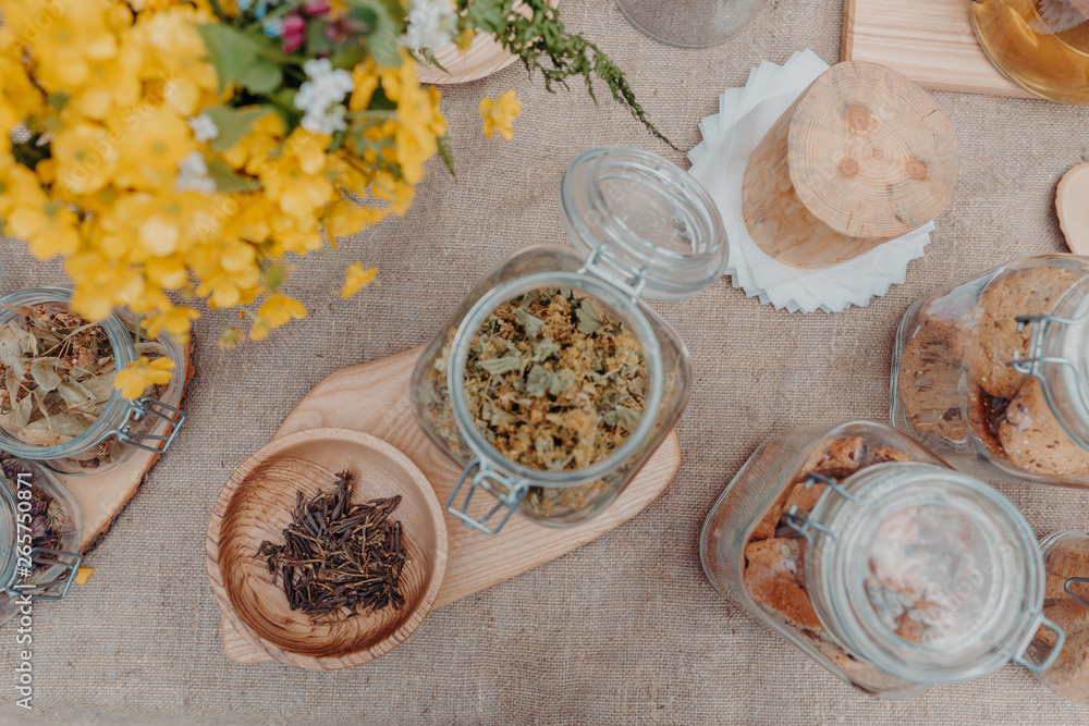 Tea herbs (lady’s mantle, sagan daylya, blooming sally or ivan chay) in bowl and jars on table with yellow flowers and jars with cookies near. Grey background. Horizontal orintation, top view. Tea herbs (lady’s mantle, sagan daylya, blooming sally or ivan chay) in bowl and jars on table with yellow flowers and jars with cookies near. Grey background. Horizontal orintation, top view.