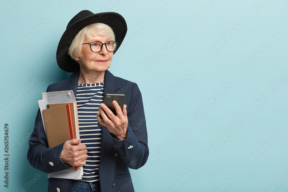 Headshot of beautiful old lady looks aside, has thoughtful expression, sends text messages in online chat, holds papers and notebook, wears stylish black hat and formal coat, stands over blue wall - obrazy, fototapety, plakaty 