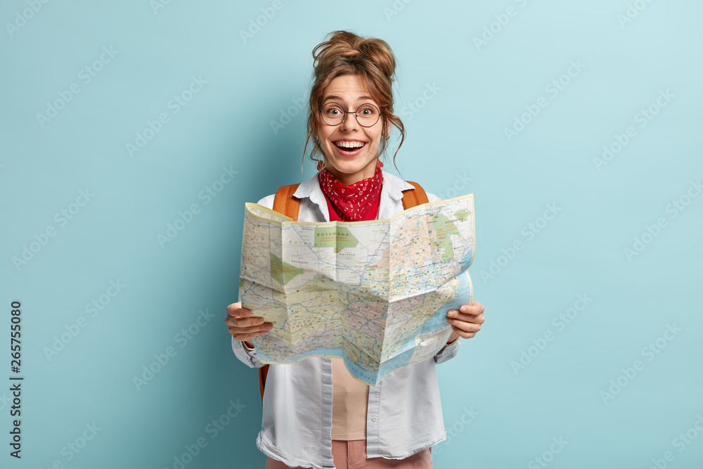 Happy female traveler holds map, checks route for destination, has trip across Europe during holidays, carries backpack, wears round spectacles, isolated on blue wall, plans tour, checks location Happy female traveler holds map, checks route for destination, has trip across Europe during holidays, carries backpack, wears round spectacles, isolated on blue wall, plans tour, checks location