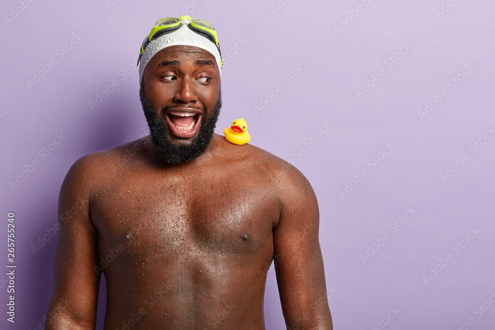 Summer holiday and recreation concept. Strong African American man with thick beard, being wet after swimming in sea or pool, has toy rubber duckling on muscular shoulder, opens mouth widely