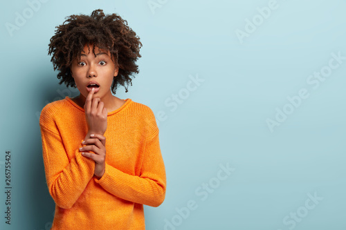 Hald length shot of impressed surprised young Afro American woman looks with bated breath, wintenesses something terrible, dressed in orange jumper, stands over blue background. Reaction concept photo