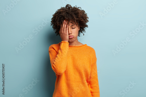 Photo of tired dark skinned female covers face, closes eyes, feels fatigue, needs good rest, dressed in orange jumper, has sleepy expression, isolated over blue background. Weariness and people