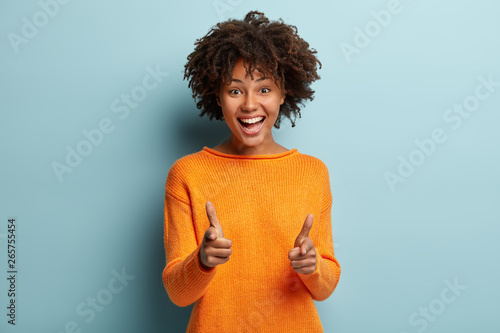 Joyous pretty African American lady makes finger gun gesture at camera, expresses choice, smiles broadly, dressed in orange jumper, isolated over blue background, says you are chosen. Bang, bang photo