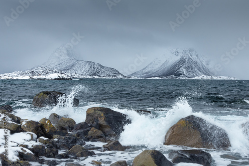 Waves crushing ont he rocks on a beach from Lofoten Islands, Norway. Spectacular snowy mountains in the background. Moody witner day photo