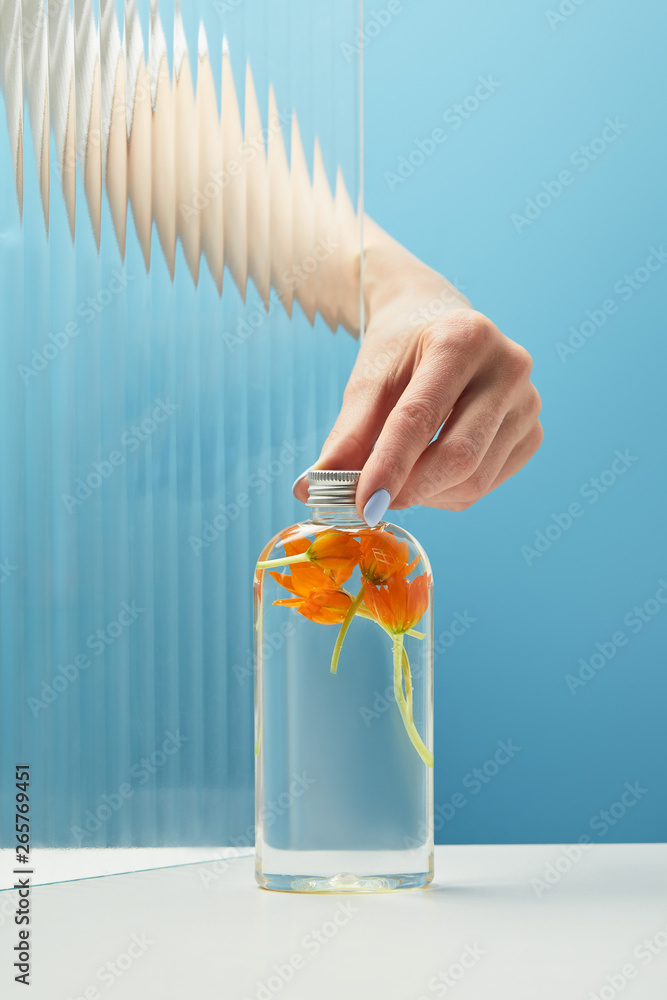 cropped view of woman putting cap on bottle with organic beauty product with orange flowers on blue background cropped view of woman putting cap on bottle with organic beauty product with orange flowers on blue background