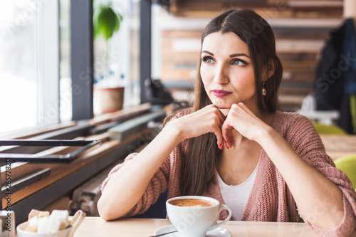 Beautiful young woman sits at a table in a cafe with a cup of coffee and looks out the window