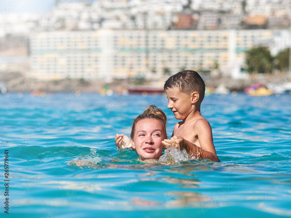 Cute European boy is splashing in the sea with his mother. They are having fun during their holidays on the seacoast.