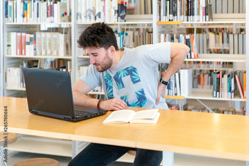 Young student with back pain from sitting so long in front of the laptop in the library Young student with back pain from sitting so long in front of the laptop in the library