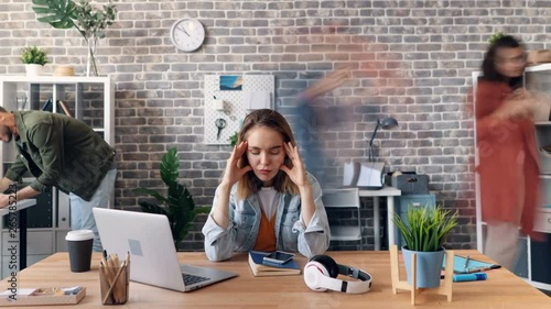 Zoom out time-lapse of tired young woman exhausted employee touching head in office feeling sick sitting at desk while busy coworkers are rushing around. photo