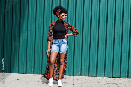 African woman with afro hair, in jeans shorts and white sunglasses posed against green steel wall.