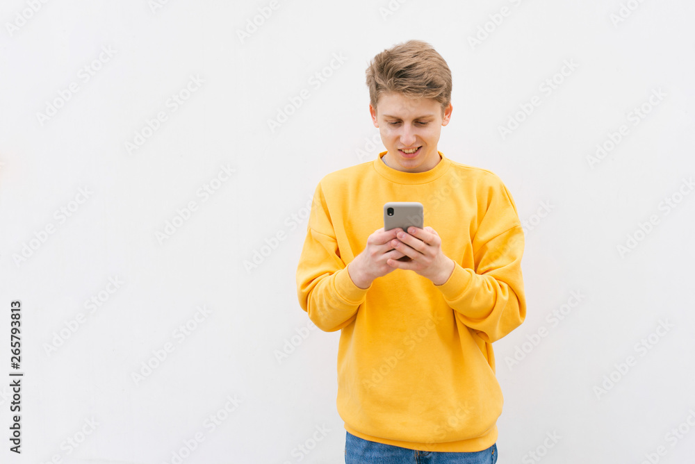 Fototapeta premium Close-up portrait of a guy in a yellow sweatshirt, standing against a white wall, enjoying a smartphone rejoicing. Happy young in casual clothing writes a message on the smartphone and smiles