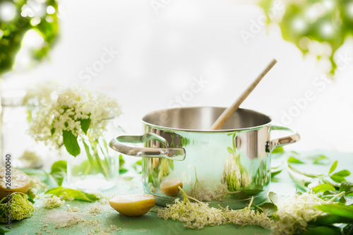 Elder flowers syrup cooking preparation. Pot with wooden spoon on kitchen table with sugar, lemon and elderflowers. Healthy seasonal natural food concept. Copy space for your recipes