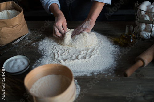 Women's hands knead the dough. Dough, flour with accessories for the kitchen in the composition on the kitchen table. Toned image. Selective focus.