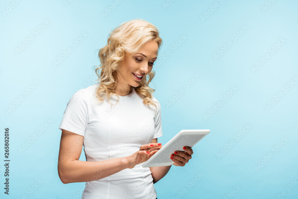 happy blonde woman using digital tablet while standing on blue