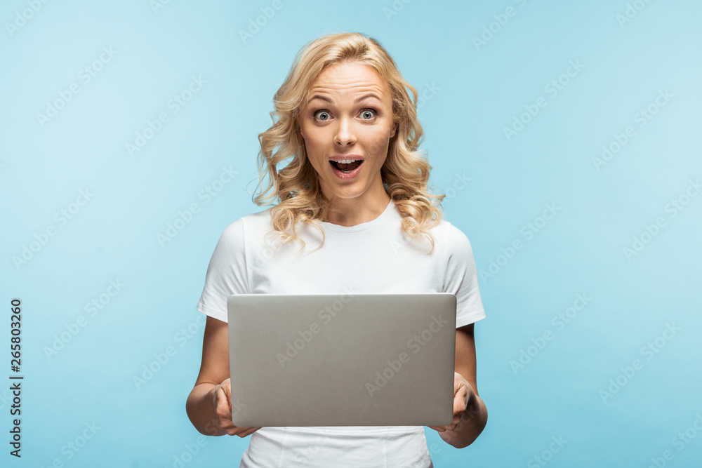Naklejka premium surprised blonde woman holding laptop and looking at camera on blue