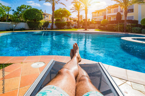 Relaxing at the swimming pool. Man relaxing next to swimming pool. Man enjoying the hot summer at swimming-pool. Sunbathing by the swimming pool, mans legs lying down on a sun lounger over the water. photo