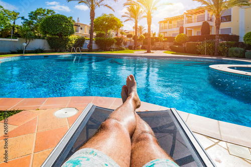 Obraz na płótnie Relaxing at the swimming pool. Man relaxing next to swimming pool. Man enjoying the hot summer at swimming-pool. Sunbathing by the swimming pool, mans legs lying down on a sun lounger over the water. w salonie
