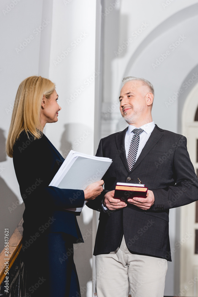 low angle view of happy mature teacher holding books and looking at attractive colleague with folder in hands low angle view of happy mature teacher holding books and looking at attractive colleague with folder in hands