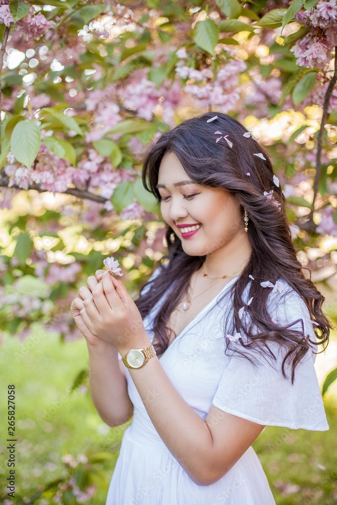 A young beautiful Asian woman in a white dress walks in a flowered park. Sakura. Blooming trees. A young beautiful Asian woman in a white dress walks in a flowered park. Sakura. Blooming trees.