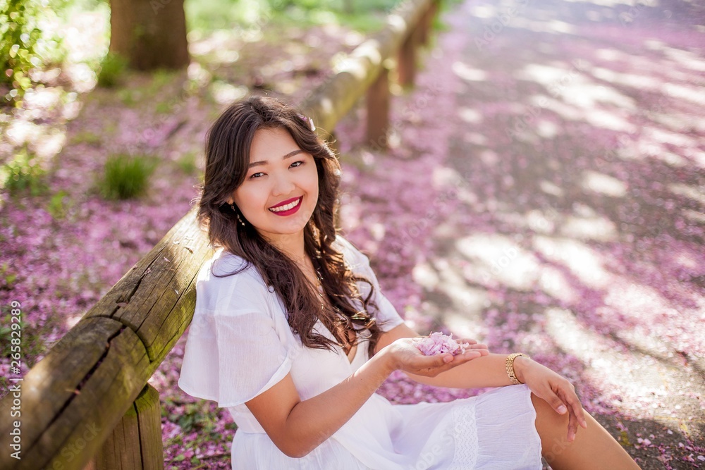 A young beautiful Asian woman in a white dress walks in a flowered park. Sakura. Blooming trees. A young beautiful Asian woman in a white dress walks in a flowered park. Sakura. Blooming trees.