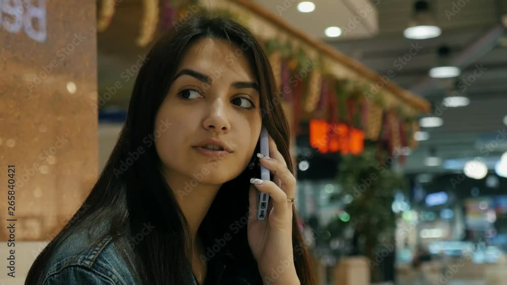 Positive cute girl in jeans jacket sitting on the bench in the mall and talking with girlfriend on the phone Positive cute girl in jeans jacket sitting on the bench in the mall and talking with girlfriend on the phone
