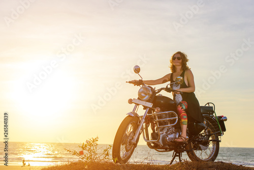A sexy young woman in sunglasses sits on a black chrome motorbike. Hipster girl on a motorcycle against the sunset. The concept of women's independence.