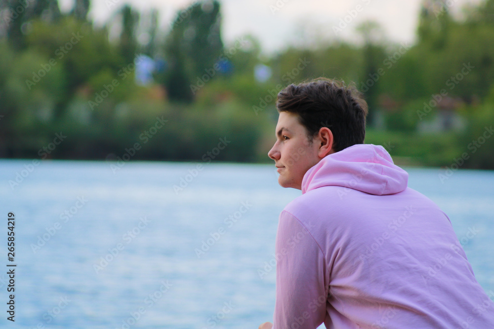 Profile of a boy with brown hair who smiles looking in front of him. Teenager in pink sweater contemplating a lake standing on a wooden bridge Profile of a boy with brown hair who smiles looking in front of him. Teenager in pink sweater contemplating a lake standing on a wooden bridge