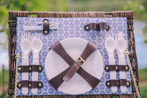 Close-up of a picnic suitcase basket with dishes - plates, spoons and forks on a blurred green tree background