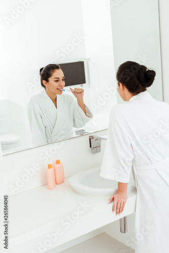 selective focus of happy young woman brushing teeth and looking at mirror in bathroom