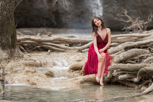 Fototapeta Naklejka Na Ścianę i Meble -  Young beautiful woman is wearing fashion red dress sitting in forest near the waterfall