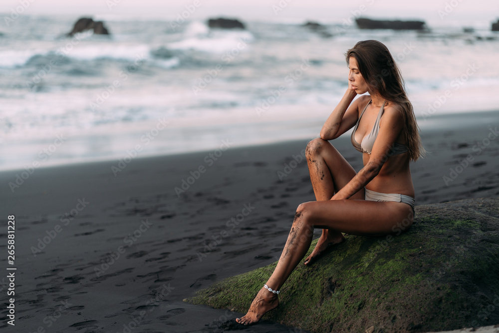 Cute sexy girl in beige swimwear rests on black volcanic sand beach. Cute sexy girl in beige swimwear rests on black volcanic sand beach.