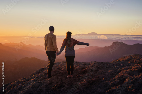 young couple from behind holding hands while standing on mountain top with scenic landscape and sunset in the background