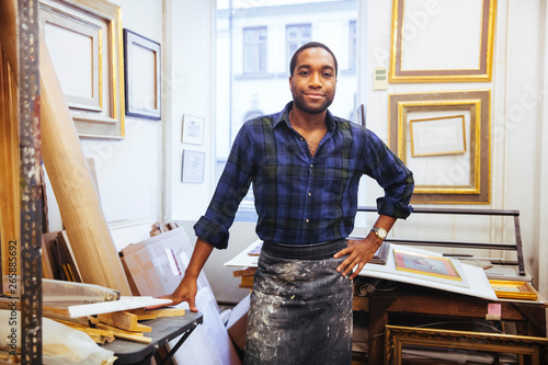 Portrait of smiling young male artist standing with hand on hip at framing workshop photo