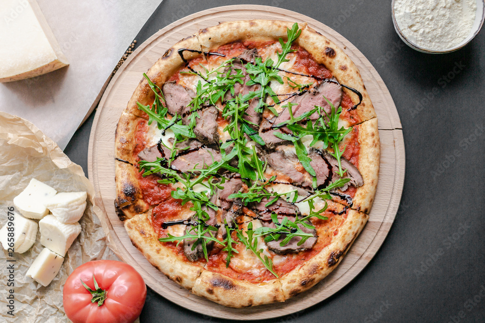 Pizza with roast beef, arugula on the wooden plate. Tomato and pieses of cheese near on the table. Horizontal. Natural light. Black background. Flat lay, top view. Pizza with roast beef, arugula on the wooden plate. Tomato and pieses of cheese near on the table. Horizontal. Natural light. Black background. Flat lay, top view.