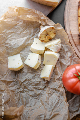 Cheese pieces with fried half of garlic and tomato on white cooking paper with tomatoe and wooden cutting board. Top view, flat lay, black background, copy space.