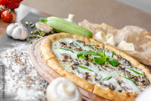 Pizza with zucchini on the wooden plate in the restaurant. Onion, zucchini, champignons, white bottle near on the table. Horizontal image. Natural light. Black background. Shallow focus.